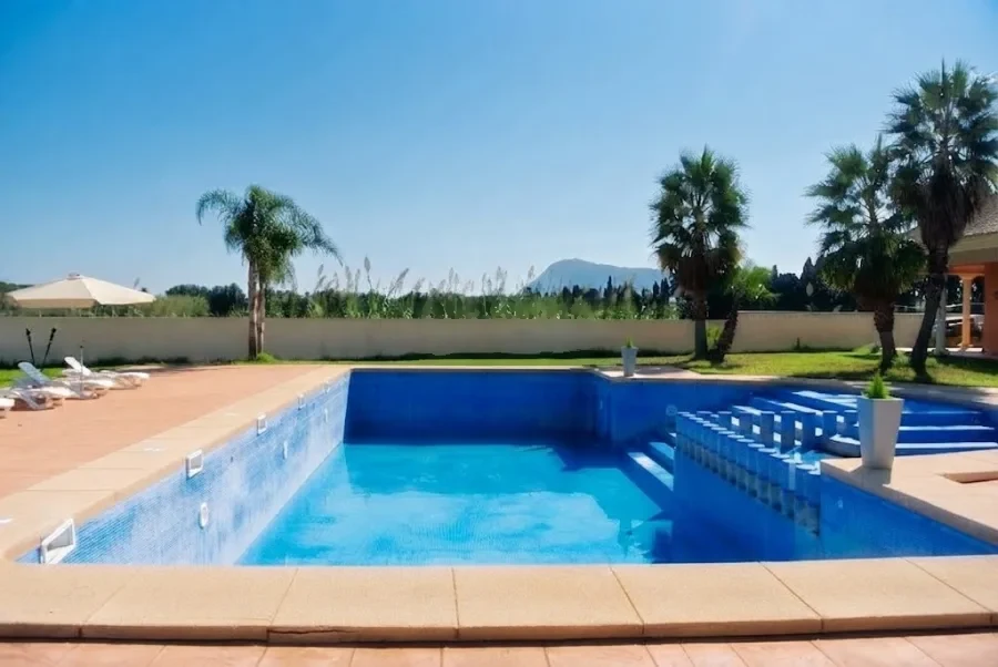 La piscina rectangular de gresite azul, con palmeras y el muro del jardín al fondo