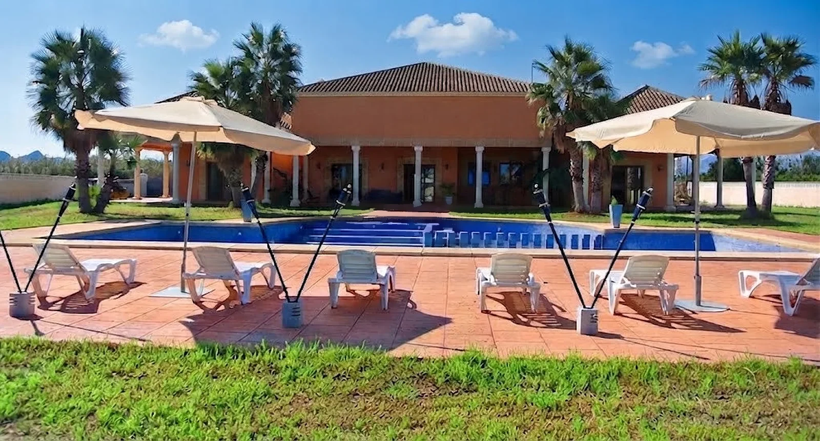 Terraza junto a la piscina con tumbonas blancas y grandes sombrillas, y la casa detrás